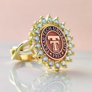 May include: A gold-toned ring featuring a circular centerpiece with the Texas Tech University logo and the year 1923. The central emblem is surrounded by a halo of small, clear, sparkling stones set in a scalloped gold-toned metal frame.