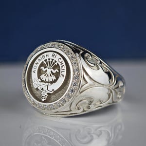Könnte beinhalten: Ein silberner Ring mit einem schwarzen und weißen Wappen, das ein stilisiertes Design eines Vogels mit Pfeilen und dem Text "Aonaibh Ri Cheile" zeigt. Der Ring ist mit klaren Zirkonia-Steinen um das Wappen herum besetzt.