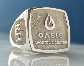 May include: A silver-toned metal ring featuring a square face with a textured background. The face displays a teardrop icon above the text OASIS and APOSTOLIC CENTER. The side of the ring is engraved with the letters RH in a bold, serif typeface.