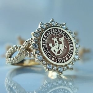 May include: A gold ring with a circular emblem inscribed with "SOUTHERN ARKANSAS UNIVERSITY MBA 1909". The ring is embellished with small, clear gemstones around the emblem and on the band. The ring is displayed on a reflective surface.