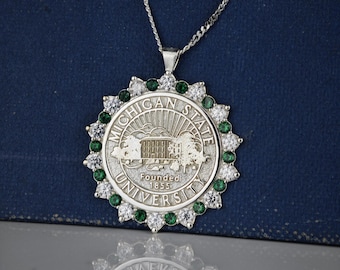 May include: A silver pendant necklace with a circular charm featuring the Michigan State University logo. The charm is surrounded by a border of clear and green gemstones.