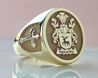 May include: A gold signet ring featuring a circular face with an engraved crest depicting a stag and the name McCarthy. The side of the ring displays a polished cross emblem set against a textured background, with a smooth, reflective gold band.