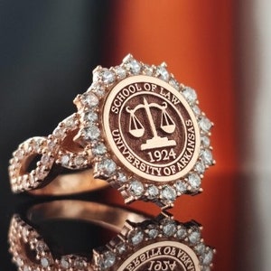 May include: A rose gold ring with a diamond-encrusted band and a circular top featuring the University of Arkansas School of Law logo. The logo includes a scale of justice, the words "School of Law", "University of Arkansas", and the year "1924".