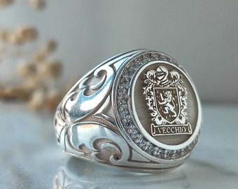 May include: A silver ring with a detailed design featuring a crest with a lion and the word "Vecchio". The crest is surrounded by a circle of small clear stones.