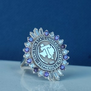 Könnte beinhalten: Ein silberner Ring mit einer Fassung aus violetten und weißen Steinen. Der Ring zeigt das Logo der Tarleton State University, das die Worte "Tarleton State University", "Stephenville, Texas" und "1899" enthält.