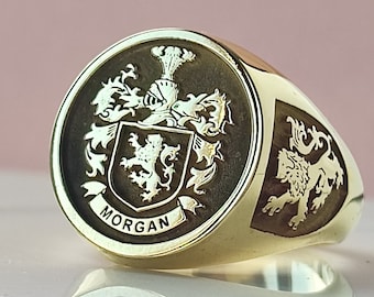 May include: A gold signet ring featuring a circular face with an engraved coat of arms, a knight's helmet, and a lion rampant. The name MORGAN is inscribed on a banner below the shield. A lion rampant is also engraved on the side of the ring band.