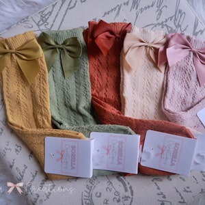 May include: A collection of knee-high socks in various colors, including mustard yellow, olive green, rust, cream, and pink. Each sock features a large satin bow at the top. The socks have a delicate, openwork design. Tags with the "Sorella Creations" logo are attached.