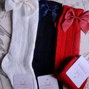 May include: Three pairs of knee-high socks with bows. The socks are white, black, and red. Each pair has a different colored bow. The socks are made by Sorella Creations.