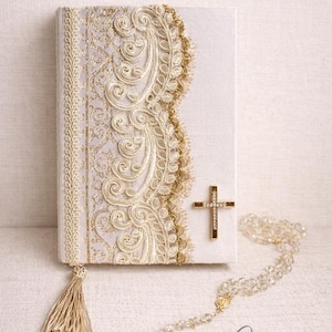 May include: A white book with gold embellishments, a gold cross, and a rosary. The book cover features intricate gold lace and a gold tassel. The cross is attached to the book, and the rosary is made of clear beads.