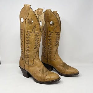 May include: A pair of tan leather cowboy boots. The boots have decorative stitching, a pointed toe, and a stacked heel. The shaft of the boots features a large cut-out circle near the top. The boots are a classic Western style.
