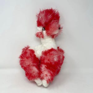 May include: A vintage plush poodle toy in a seated position. The poodle has a white body with red and white fluffy fur on its head and legs. It has black eyes and a small, pink nose. The toy is set against a plain white background.