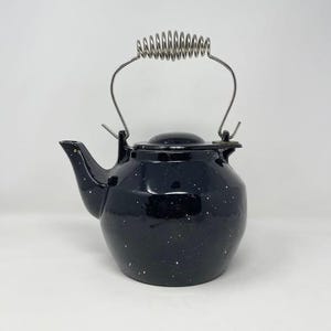 May include: A black enamel teapot with a speckled white pattern. It features a curved metal handle with a coiled grip and a matching lid. The spout is curved, and the teapot has a classic, rounded shape.