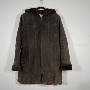 May include: A brown suede coat with a hood and a full-length zipper. The coat has a fur-lined hood and cuffs. The coat is hanging on a black hanger against a white background. The coat is a medium length.