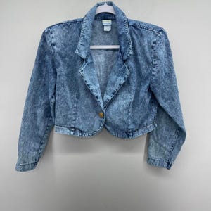 May include: A cropped, acid-wash denim jacket with long sleeves and a single gold button closure. The jacket features a notched collar and is hanging on a white hanger against a plain background. The denim has a blue and white mottled pattern.