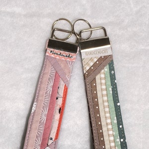 Könnte beinhalten: Zwei Stoffschlüsselanhänger mit silbernen Schlüsselringen. Der Schlüsselanhänger links ist rosa und weiß mit dem Wort "Handmade" auf dem Metallverschluss. Der Schlüsselanhänger rechts ist braun, beige und grün mit dem Wort "Handgemacht" auf dem Metallverschluss.