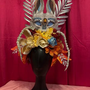 May include: A whimsical headpiece featuring a silver bunny mask with blue eyes and rhinestones, surrounded by a crown of colorful artificial flowers, including yellow, orange, red, and blue.