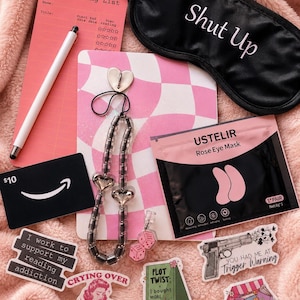 May include: A collection of items on a pink fluffy surface. Includes a black eye mask with "Shut Up" text, a reading list, a pen, stickers, a gift card, a beaded necklace, and eye masks. The stickers have various texts and graphics.