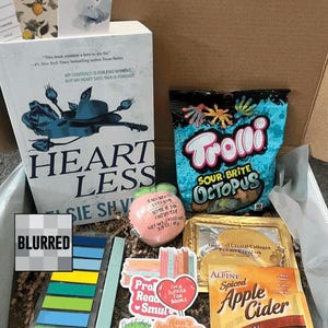 May include: A gift box filled with a book titled "Heartless", a bag of Trolli Sour Brite Octopus candies, a peach-shaped bath bomb, stickers, and a packet of Alpine Spiced Apple Cider. The box also contains a gold-framed eye mask.