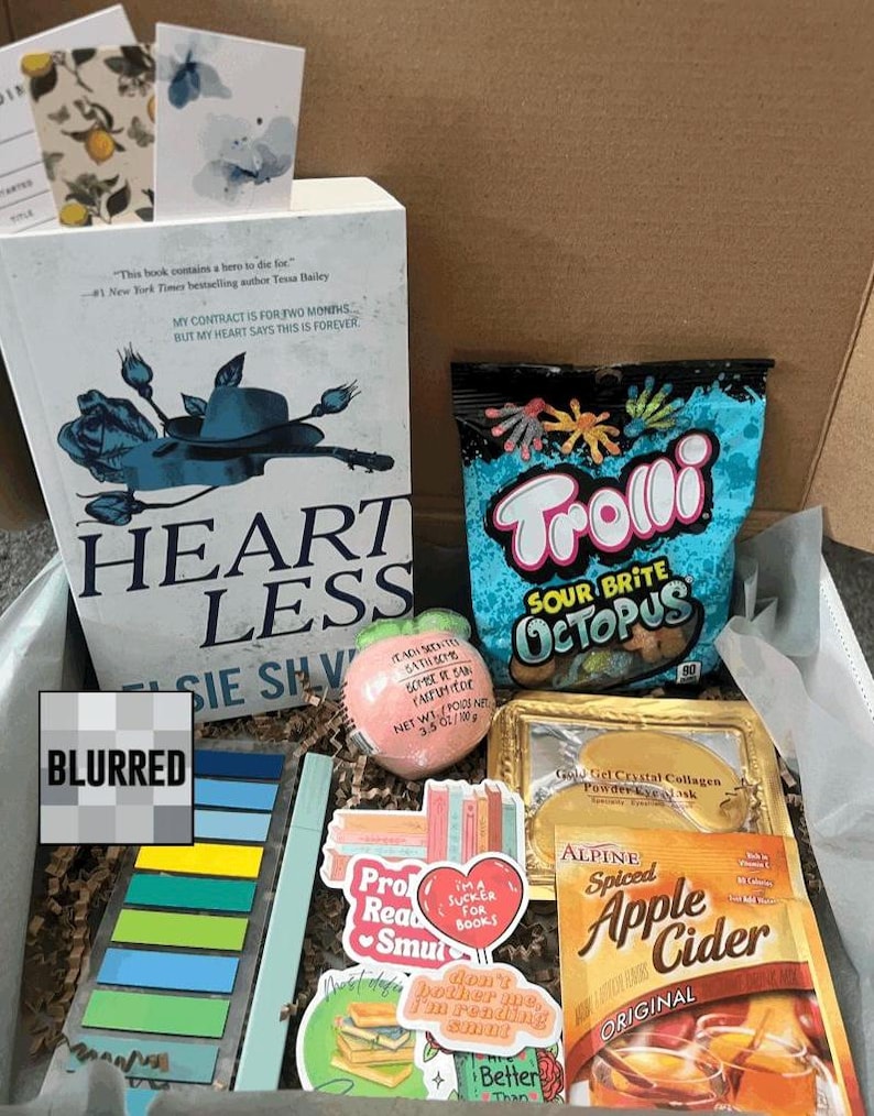 May include: A gift box filled with a book titled "Heartless", a bag of Trolli Sour Brite Octopus candies, a peach-shaped bath bomb, stickers, and a packet of Alpine Spiced Apple Cider. The box also contains a gold-framed eye mask.