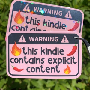 Peut inclure: Deux autocollants d'avertissement roses avec du texte noir et un graphique de piment rouge. L'autocollant du haut dit "ATTENTION ! Cette liseuse contient du contenu explicite". L'autocollant du bas dit "ATTENTION ! Cette liseuse contient du contenu explicite".