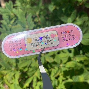 May include: Pink plaster sticker with glitter and the text "Healing Takes Time" with a smiley face and a heart.