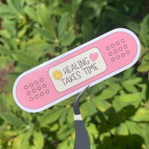 May include: Pink plaster sticker with a smiley face, a pink heart, and the text "HEALING TAKES TIME".