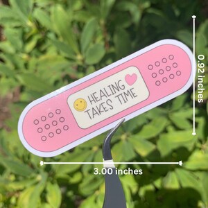 May include: Pink and white band-aid sticker with a smiley face, a heart, and the text "Healing Takes Time". The sticker is 7.62 cm long and 2.34 cm wide.