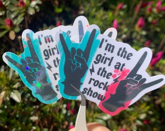 May include: Three stickers with a hand making the rock and roll hand gesture. The stickers have a holographic finish and say "I'm the girl at the rock show".
