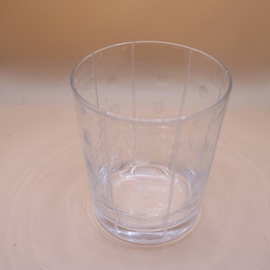 Puede incluir: Un vaso transparente de vidrio con un patrón texturizado de líneas verticales y pequeños círculos. El vaso está vacío y se encuentra sobre una superficie de color marrón claro.