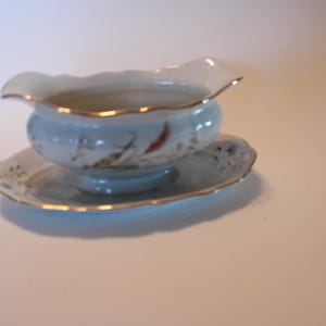 May include: A white porcelain gravy boat with a floral design and gold trim. The boat is sitting on a matching saucer.