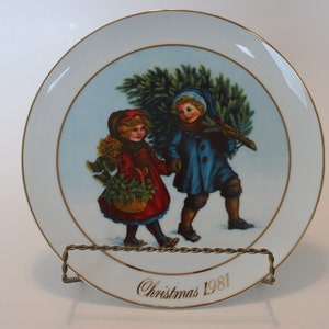 Pode incluir: Um prato de porcelana branca com borda dourada apresenta uma cena de Natal com duas crianças carregando uma árvore de Natal. O prato tem uma inscrição dourada que diz "Natal 1981".