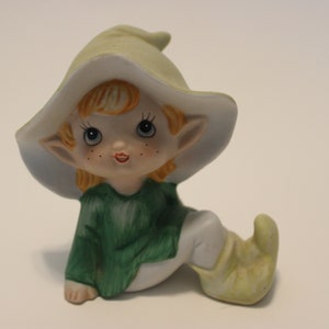 May include: A ceramic figurine of a sitting elf wearing a green and white hat, a green shirt, and white trousers. The elf has blonde hair, blue eyes, and rosy cheeks.