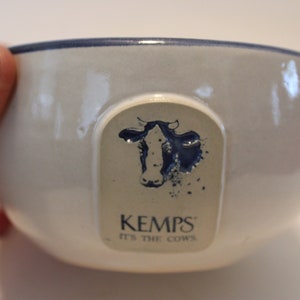 Puede incluir: Un tazón de cerámica blanco con un borde azul y una ilustración azul y blanca de una vaca con el texto "Kemps' It's the Cows" en la parte delantera.