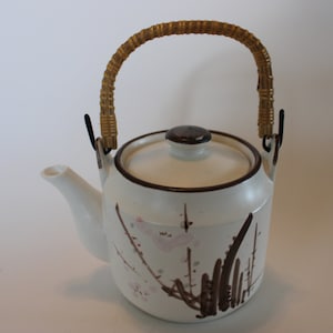 May include: White ceramic teapot with a brown and pink floral design. The teapot has a brown lid and a woven bamboo handle.