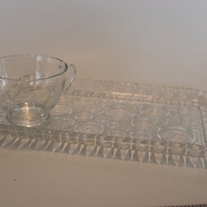 Puede incluir: Una bandeja de servir de vidrio transparente con un borde elevado texturizado y una taza pequeña de vidrio transparente con asa. La bandeja tiene un patrón de pequeños círculos.
