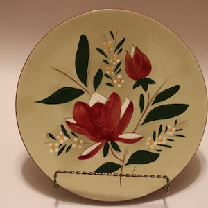 Puede incluir: Un plato redondo pintado a mano con un fondo verde claro y un borde marrón oscuro. El plato presenta un diseño floral con flores rojas y blancas, hojas verdes oscuras y pequeños detalles blancos y amarillos. El plato se exhibe en un soporte metálico.