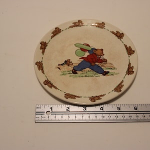 May include: A white ceramic plate with a brown teddy bear carrying balloons. The plate has a border of brown teddy bears running around the edge.