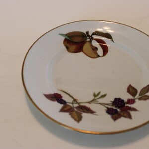May include: A white ceramic saucer with a gold rim features a painted design of red apples, green leaves, and purple berries.