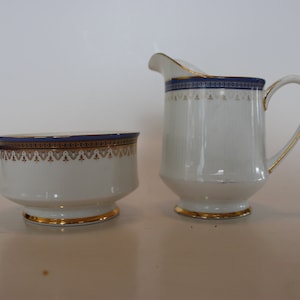 May include: A white creamer and sugar bowl set with gold trim and blue and gold accents.