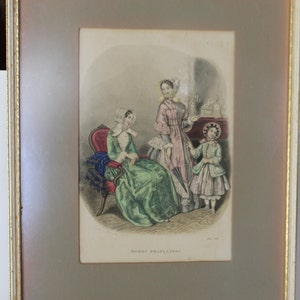 May include: A framed print depicting three women in 19th-century attire. The central figure wears a pink dress and holds a parasol. The woman on the left is seated in a chair, wearing a green dress. The woman on the right is dressed in white and carries a small basket. The print is titled "Modes Francaises".
