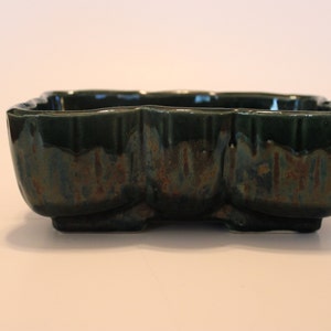 May include: A dark green ceramic planter with a textured, uneven glaze. The planter has a rectangular shape with rounded edges and a slightly flared lip.