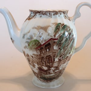 May include: A white ceramic teapot with a brown and green floral design. The teapot has a curved spout and a handle. The design features a house with a red roof and palm trees.