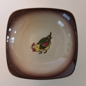 May include: A small, square, brown and white ceramic dish with a brown rim. The dish has a yellow chicken with green and red accents painted on the inside.
