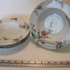 Puede incluir: Cuencos de sopa de porcelana blanca con un estampado floral de rosas rosas y amarillas y hojas verdes. Los cuencos tienen un borde dorado y se exhiben sobre una superficie blanca. Un cuenco está sobre un soporte de metal dorado.