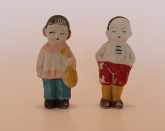 May include: Two porcelain figurines of children wearing traditional Chinese clothing. The figurine on the left is holding a yellow bag and is wearing a pink, white, and blue outfit. The figurine on the right is wearing a red and yellow outfit.