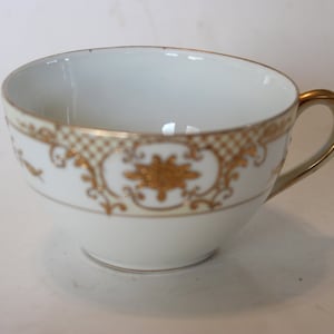 May include: A white teacup with a gold rim and intricate gold floral design. The cup has a rounded shape and a small handle.