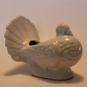 May include: White ceramic dove figurine with a detailed tail feather design. The dove is in a sitting position with its head tilted to the side. The figurine has a small opening in the back, suggesting it may be a planter.