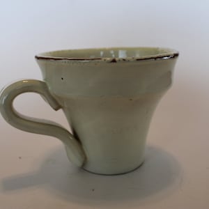 May include: A white ceramic coffee cup with a brown rim and a handle. The cup has a slightly flared shape.