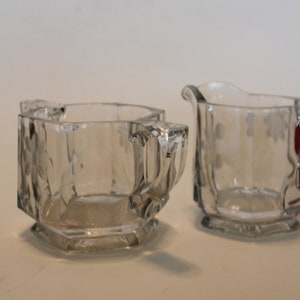May include: A set of two clear glass sugar and creamer containers with etched floral designs. The containers have octagonal bases and are made of clear glass.