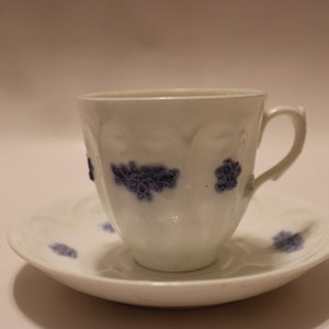 Może przedstawiać: Biały porcelanowy kubek do herbaty i spodek z niebieskim wzorem kwiatowym. Kubek ma żebrowany wzór i uchwyt. Spodek ma falisty brzeg.